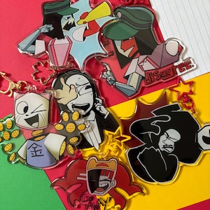 Puede incluir: Una colección de llaveros acrílicos coloridos con personajes de dibujos animados. Los llaveros muestran varios diseños, incluyendo un gato, una persona con traje y personajes con bocadillos. El fondo es una mezcla de rojo, verde y amarillo.