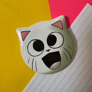 May include: A white cat-shaped badge with a large, open mouth and wide eyes. The cat has black outlines, pink inner ears, and whiskers. The background is yellow, pink, and lined paper.