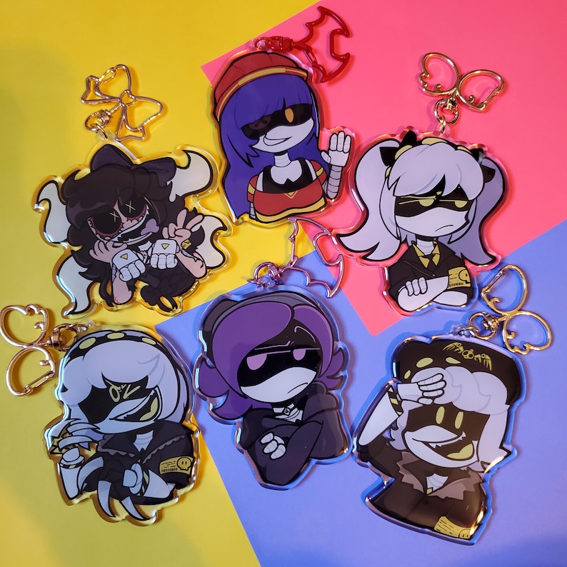 May include: Six colorful cartoon character keychains with black outlines and yellow eyes. Each keychain features a different character with a unique hairstyle and clothing. The keychains are made of clear acrylic and have gold metal rings.