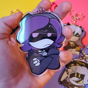 May include: A purple and black cartoon character with a serious expression, wearing a black hooded jacket, is featured on a clear acrylic keychain. The character has purple hair and a white face with black eyes.