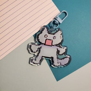 May include: A light blue keychain with a pixelated cat design. The cat has a pink mouth and a white body. The keychain is attached to a silver metal ring.