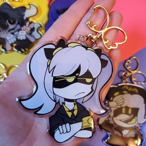 May include: A clear acrylic keychain featuring a cartoon character with purple hair, black eyes, and a yellow and black outfit. The character is wearing a black mask with yellow eyes and has their arms crossed. The keychain has a gold metal clasp.