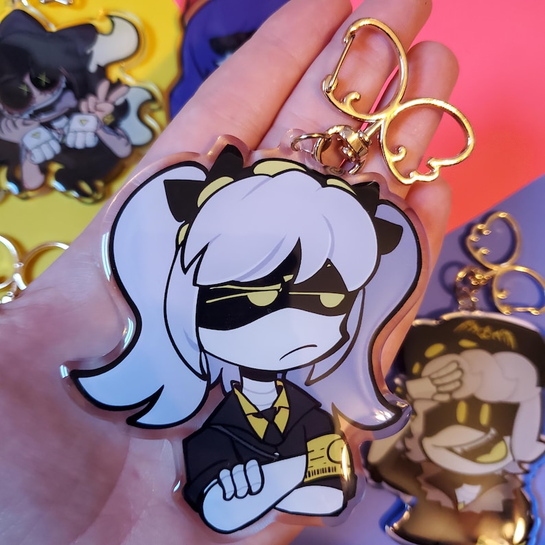 May include: A clear acrylic keychain featuring a cartoon character with purple hair, black eyes, and a yellow and black outfit. The character is wearing a black mask with yellow eyes and has their arms crossed. The keychain has a gold metal clasp.