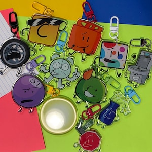 May include: A collection of acrylic keychains featuring cartoon characters with various designs and colors. Each keychain has a metal clasp for attachment. The keychains are on a bright green and yellow background.