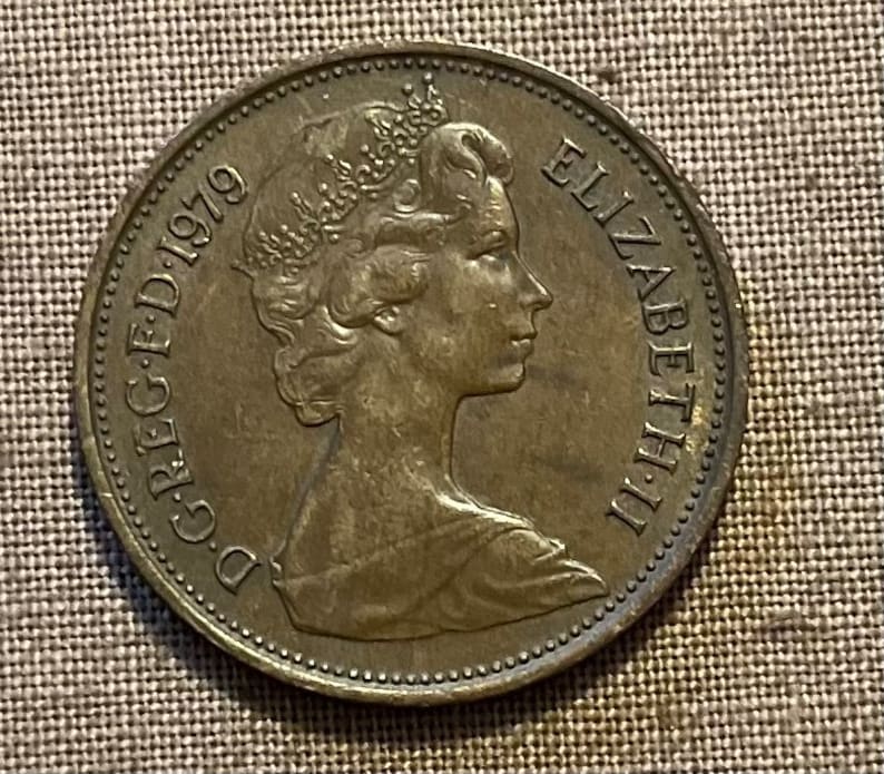 May include: A bronze-coloured British coin featuring the profile of Queen Elizabeth II with the inscription "D.G. REG. F.D. 1979" and "ELIZABETH II" on the coin.