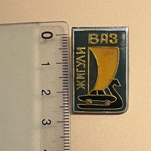 Puede incluir: Un pin de metal con fondo verde y un coche dorado con una vela. El texto "ВАЗ" e "ИЖК" es visible en el pin.