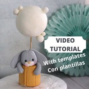 May include: A whimsical cake topper featuring a gray bunny peeking over a yellow log, topped with a white sphere with small white accents. The text "VIDEO TUTORIAL" is visible.