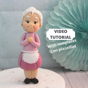 Puede incluir: Una figurita de fondant rosa de una mujer mayor con cabello blanco, vistiendo un vestido rosa con un delantal blanco y zapatos negros. La figurita está de pie sobre una base de pastel blanco. El texto "VIDEO TUTORIAL With templates Con plantillas" es visible en la imagen.