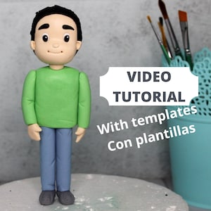 May include: A green and blue fondant figure of a person with a smiling face. The figure is standing on a white surface. The text "VIDEO TUTORIAL" is in a white box with rounded corners. The text "With templates" and "Con plantillas" is below the box.