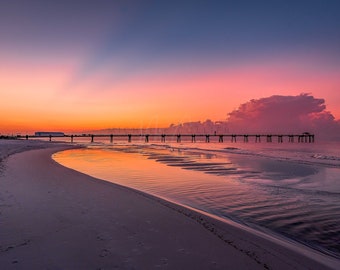 May include: A long wooden pier extends out into the ocean at sunset. The sky is a vibrant mix of pink, orange, and purple, reflecting in the calm water. The beach is sandy and empty, with a few footprints in the sand.