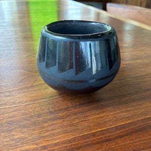 May include: A small, rounded black ceramic bowl with a polished finish. The exterior features a matte black geometric pattern consisting of vertical lines and triangular shapes encircling the upper portion of the vessel, creating a contrast.