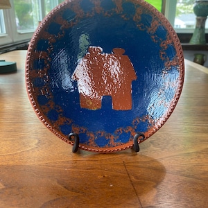 May include: A handmade ceramic plate with a blue interior and a brown house design. The rim features a decorative brown pattern. The plate is displayed on a black metal stand, showcasing its unique craftsmanship and design.