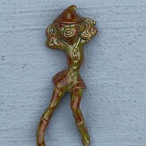 May include: A ceramic figurine of a girl wearing a red hat and a green dress. The figurine is glazed in a brown and green color scheme.