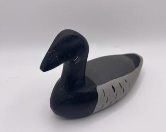 Puede incluir: Un señuelo de pato de madera tallado a mano, pintado en negro y gris. El pato tiene un cuello largo y un cuerpo redondeado.
