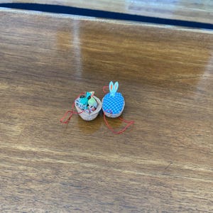 May include: Two handmade Easter-themed ornaments. One is a small basket filled with colorful miniature items. The other is a blue and white checkered bunny with long ears. Both are attached to red strings.