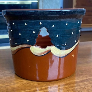 May include: A ceramic candle holder with a Christmas tree cutout. The holder is painted with a dark blue sky design with white dots, a brown base, and yellow accents. The candle holder is a decorative item.