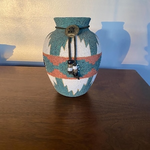 May include: A decorative vase with a Southwestern design. The vase features a turquoise, white, and coral pattern. A leather cord with a decorative button and beads is wrapped around the neck of the vase. The vase is approximately 6 inches tall.