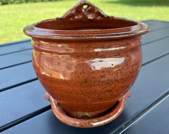 May include: A handcrafted, reddish-brown ceramic wall planter with a decorative top and attached saucer. The planter has a glossy finish and is designed for displaying small plants or flowers. The planter is sitting on a black table.