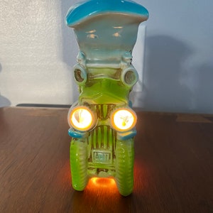 May include: A vintage ceramic car-shaped lamp in shades of green and blue. The headlights are illuminated, casting a warm glow. The lamp has a classic car design with a hat-like top. The overall aesthetic is retro and whimsical.