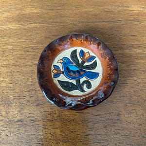 May include: A small, round ceramic dish with a brown rim and a central design. The design features a blue bird with a flower and leaf motif. The dish has a glossy finish and is likely handmade.