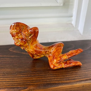 May include: A ceramic figurine of a reclining figure with arms behind the head. The figurine is glazed in shades of brown and yellow, with details in the hair and clothing. The figure is posed on a dark wooden surface.