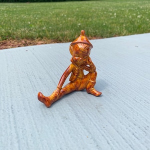 May include: A ceramic figurine of a sitting elf with a pointed hat. The elf is wearing a brown and orange outfit and has a thoughtful expression on its face.