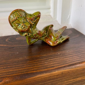 May include: A green ceramic figurine of a girl with red accents, lying on her side with her head resting on her hand. She is wearing a hat and a dress.