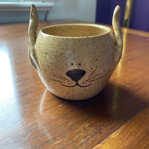 May include: A speckled ceramic mug with a cat face design. The mug is tan with black details for the eyes, nose, and whiskers. Two curved, ear-shaped handles extend upwards from the rim. The mug sits on a wooden surface.