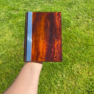 May include: A rectangular, brown and orange decorative box with a glossy finish. The box has a silver-colored metal strip down the spine and a dark gray spine. The box is held against a background of green grass.