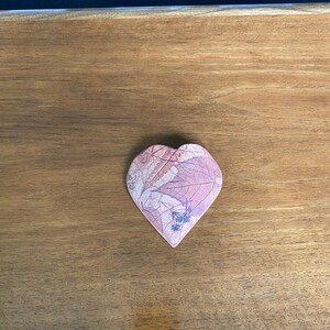 May include: A heart-shaped coaster with a pink leaf pattern. The coaster is made of a soft material and is placed on a wooden surface. The coaster has a light pink and white color scheme.