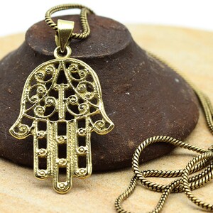Può includere: Una collana dorata con un grande ciondolo a forma di mano di Hamsa decorato. Il ciondolo è realizzato con un lavoro di filigrana intricato e presenta un design dettagliato.