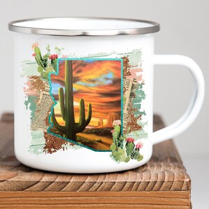 May include: White enamel mug featuring a Southwestern desert landscape design. The design includes a saguaro cactus, a sunset, and the outline of Arizona. The mug has a silver rim and a white handle.