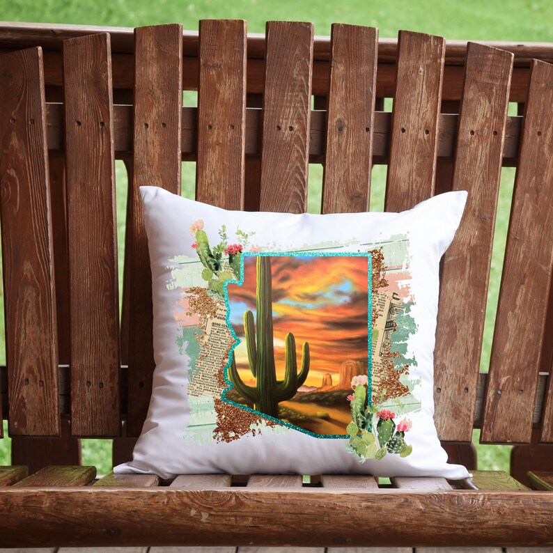 May include: White square pillow featuring a desert landscape design. The design includes a large green cactus, a sunset, and a turquoise outline of the state of Arizona. The pillow is decorated with cactus and floral accents.