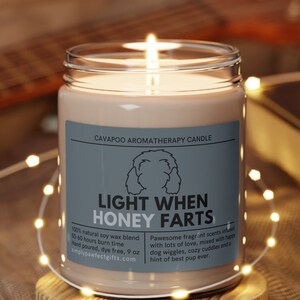 May include: A light brown aromatherapy candle with a white label that reads "CAVAPOO AROMATHERAPY CANDLE" and "LIGHT WHEN HONEY FARTS". The candle is on a wooden coaster and surrounded by white fairy lights.