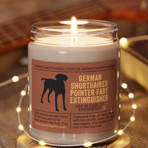 May include: A brown glass candle with a white label that reads "German Shorthaired Pointer Fart Extinguisher" and "Use in Case of Emergency". The candle is on a wooden coaster and surrounded by white string lights.