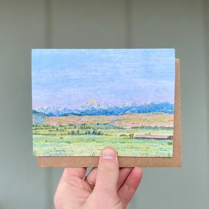 Op de afbeelding: Een kaart met een pasteltekening van een bergketen met een klein gebouw op de voorgrond. De lucht is lichtblauw met een paar wolken. De bergen zijn lichtpaars en lichtblauw. Het gebouw is donkerbruin.