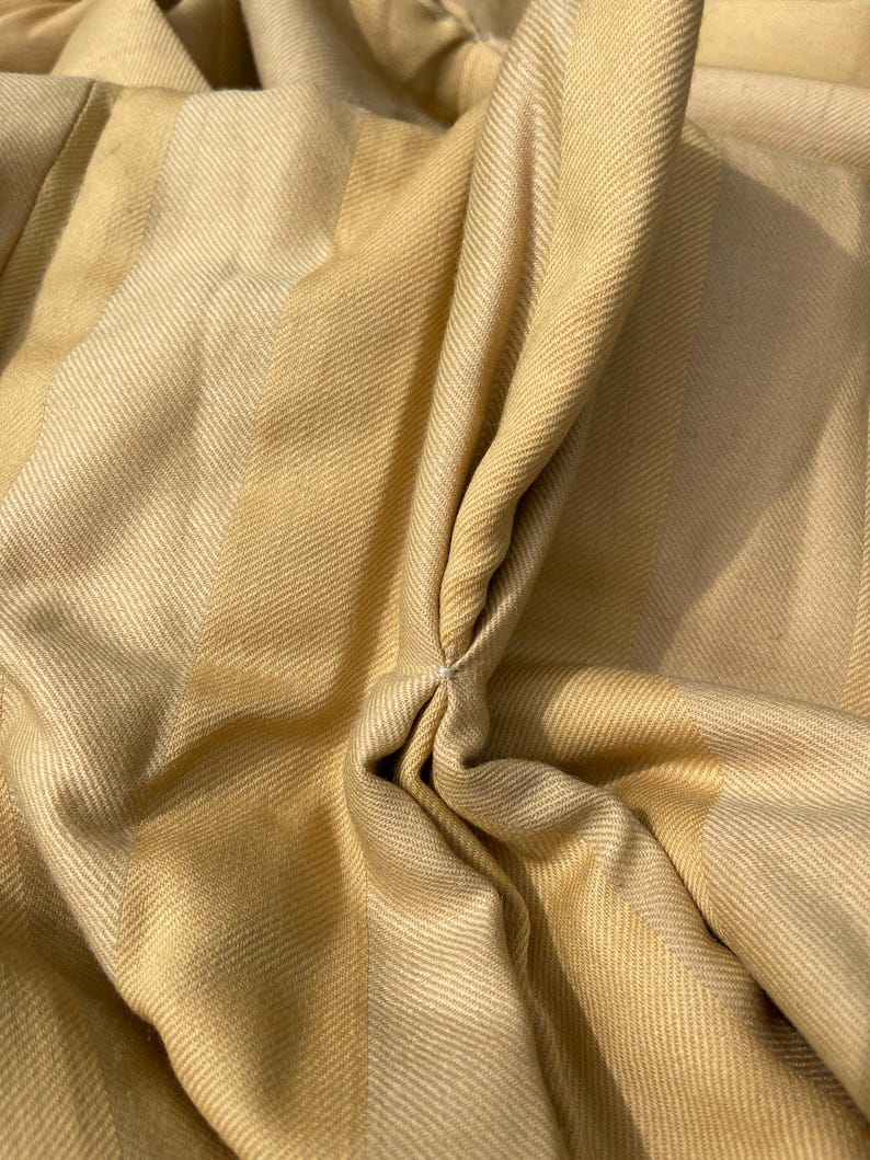 May include: Close-up of a beige and light brown striped fabric with a textured weave. The fabric is folded and gathered in the center, creating a soft, flowing look.