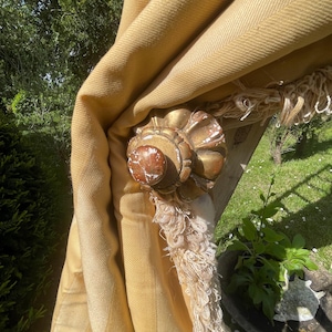 May include: A gold-painted wooden finial with a decorative design. The finial is attached to a yellow fabric curtain with a fringe trim.