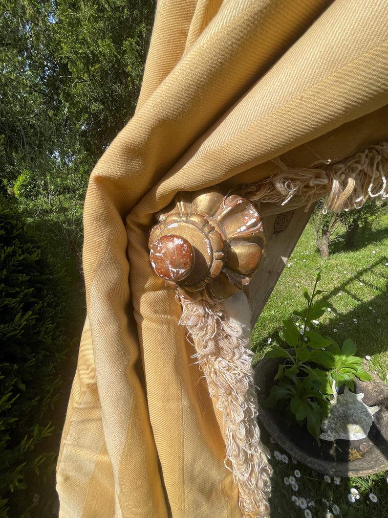 May include: A gold-painted wooden finial with a decorative design. The finial is attached to a yellow fabric curtain with a fringe trim.