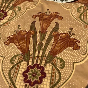 Puede incluir: Una tela beige con un patr&oacute;n repetido de lirios naranjas estilizados y un motivo floral verde, marr&oacute;n y blanco. La tela tiene una textura sutilmente punteada.