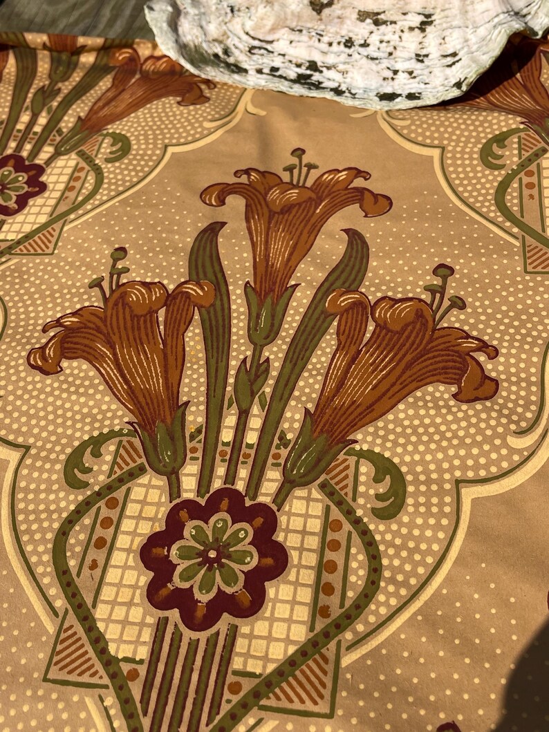 Puede incluir: Una tela beige con un patr&oacute;n repetido de lirios naranjas estilizados y un motivo floral verde, marr&oacute;n y blanco. La tela tiene una textura sutilmente punteada.