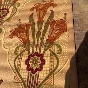 Puede incluir: Una muestra de papel tapiz vintage con un patr&oacute;n repetitivo de lirios naranjas, hojas verdes y un motivo floral burdeos sobre un fondo beige con un patr&oacute;n de puntos blancos.