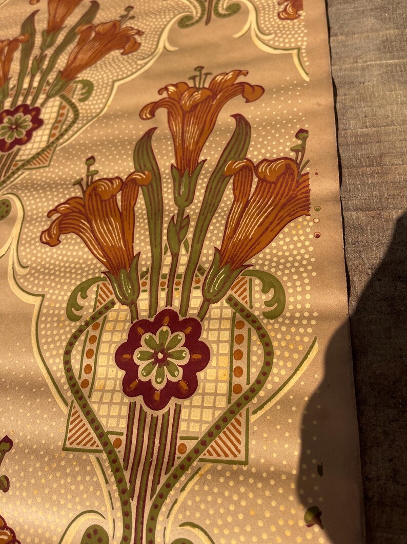 Puede incluir: Una muestra de papel tapiz vintage con un patr&oacute;n repetitivo de lirios naranjas, hojas verdes y un motivo floral burdeos sobre un fondo beige con un patr&oacute;n de puntos blancos.