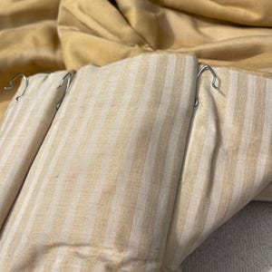 May include: Close-up of a pair of beige and white striped curtains with metal hooks attached. The curtains are folded and the fabric is textured.
