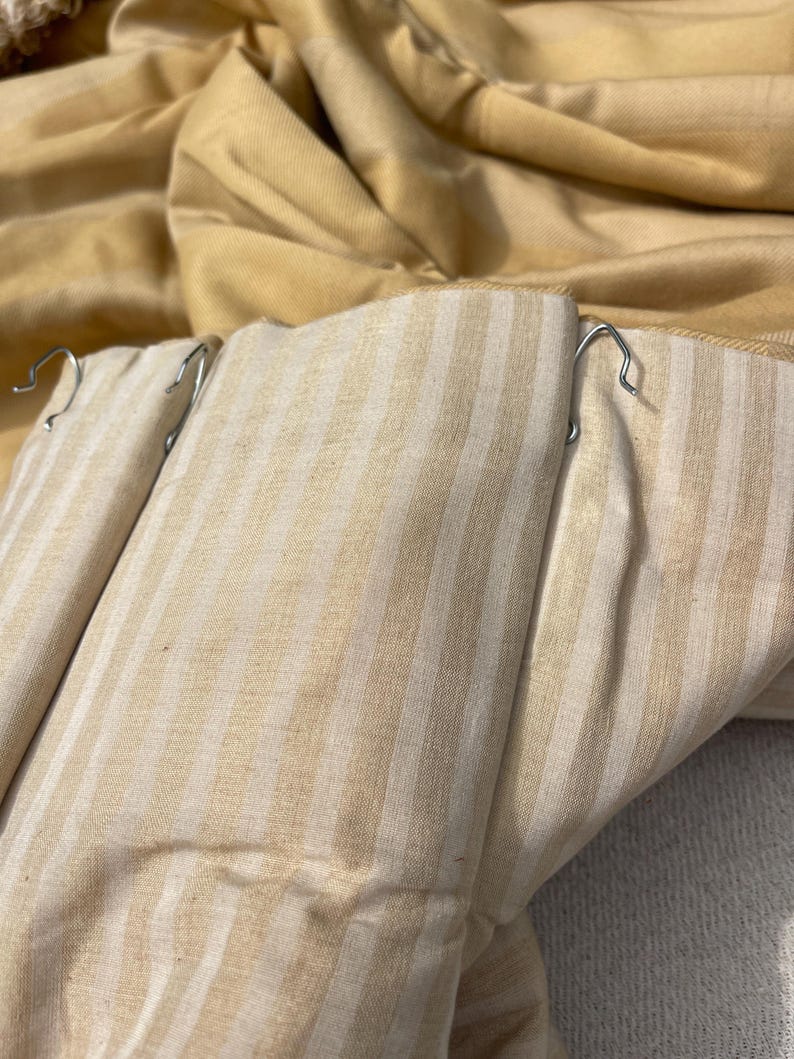 May include: Close-up of a pair of beige and white striped curtains with metal hooks attached. The curtains are folded and the fabric is textured.