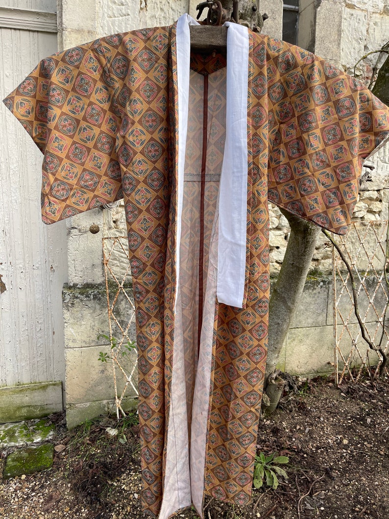 Puede incluir: Un kimono largo marr&oacute;n y dorado con un patr&oacute;n geom&eacute;trico. El kimono tiene una faja blanca y est&aacute; colgado en una percha de madera.