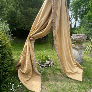 May include: A pair of long, golden yellow curtains with a fringe trim. The curtains are draped over a wooden pole and are hanging in a garden setting.