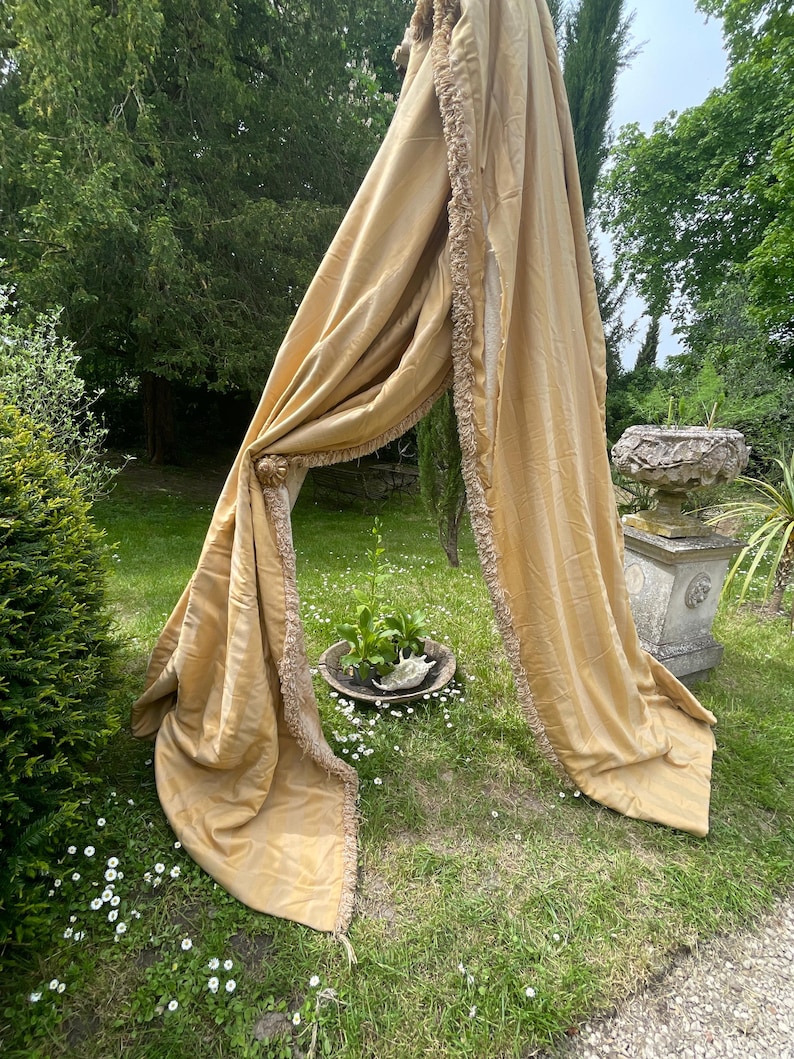 May include: A pair of long, golden yellow curtains with a fringe trim. The curtains are draped over a wooden pole and are hanging in a garden setting.