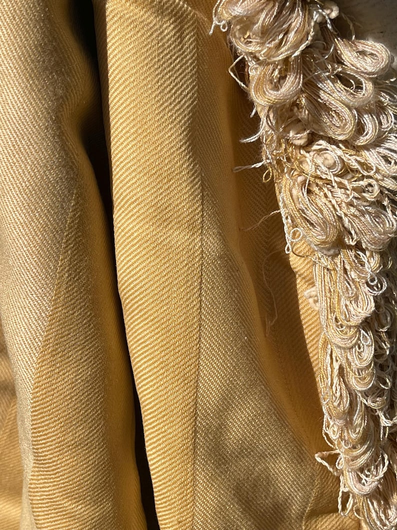 May include: A close-up of a mustard yellow woven throw blanket with a fringe border. The blanket has a herringbone pattern.