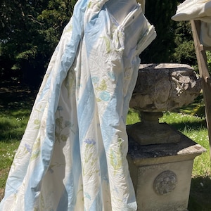 May include: A blue and white floral patterned quilt with blue vertical stripes. The quilt is hanging outdoors, with a stone pedestal and greenery in the background. The quilt features a delicate floral design.